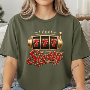 May include: An olive green short-sleeve t-shirt featuring a graphic of a gold slot machine with the number 777 displayed on the reels. Above the slot machine is the text I FEEL and below it is the word Slotty in a large red cursive font.