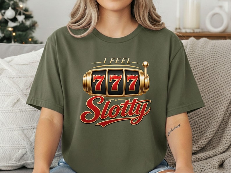 May include: An olive green short-sleeve t-shirt featuring a graphic of a gold slot machine with the number 777 displayed on the reels. Above the slot machine is the text I FEEL and below it is the word Slotty in a large red cursive font.