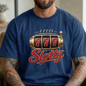 May include: A navy blue short-sleeve t-shirt featuring a graphic of a gold slot machine reel displaying the number 777. Above the graphic is the text I FEEL and below the graphic is the word Slotty in a stylized red and gold font.