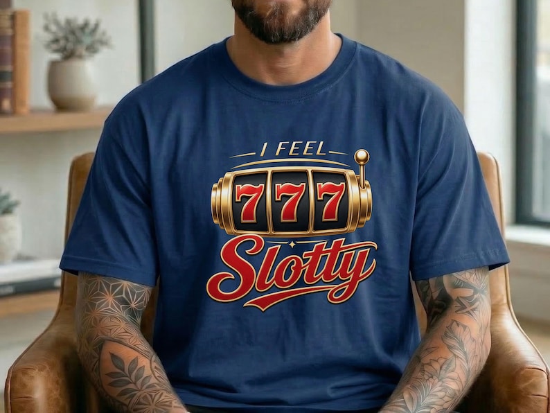 May include: A navy blue short-sleeve t-shirt featuring a graphic of a gold slot machine reel displaying the number 777. Above the graphic is the text I FEEL and below the graphic is the word Slotty in a stylized red and gold font.