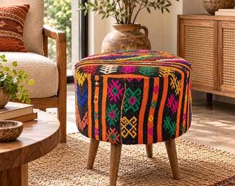 Può includere: Un pouf rotondo caratterizzato da un vivace tessuto intrecciato con motivi geometrici in nero, arancione, rosa, verde, giallo e blu. Il complemento d'arredo è sostenuto da quattro gambe in legno affusolate che offrono una base stabile.