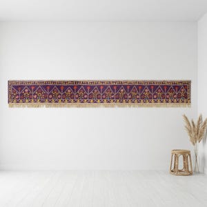 May include: A long, rectangular, purple textile with a repeating floral pattern in red, yellow, and blue. The textile has a beige fringe along the bottom edge. The textile is hanging on a white wall.