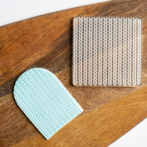 May include: Two silicone baking mats on a wooden chopping board. One mat is a square with a white knit pattern. The other mat is a light blue arch shape with a knit pattern.