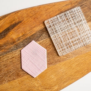May include: Two textured coasters, one pink and one clear, on a wooden surface. The pink coaster is hexagonal and the clear coaster is square.