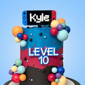 May include: A two-tier birthday cake decorated with colourful balls and a Nintendo Switch themed topper. The cake is frosted in blue, red, and black with the words "Level 10" and "Kyle" on the topper.