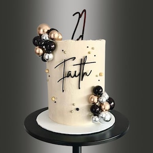 May include: A beige cake decorated with black, gold, and silver round ornaments. The cake is topped with a black acrylic topper that says "Faith" and a black acrylic number "21" topper.