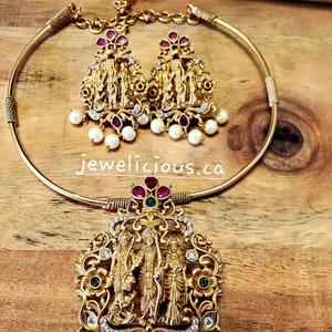 May include: A gold-toned choker necklace set with a pendant featuring a detailed depiction of a group of figures. The pendant is adorned with white stones and green and red accents. The set also includes matching earrings with a similar design.