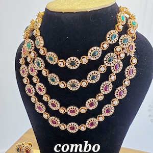 May include: A gold necklace set with multiple strands of oval-shaped stones. The stones are clear, green, and pink. The set includes matching earrings with the same stones. The text "combo" is printed on the black display stand.