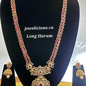 May include: A gold necklace with a long, two-strand design featuring red oval stones. The necklace has a large pendant with a peacock design and white stones. The pendant is attached to a smaller, round pendant with white stones. The necklace comes with matching earrings with red stones and white stones.
