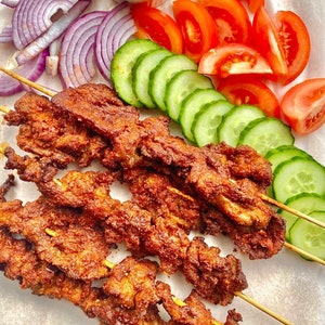 May include: A close-up of skewers of grilled meat, with a brown, crispy exterior. The skewers are arranged on a white surface, with slices of red tomatoes and green cucumbers surrounding them.
