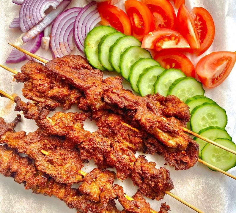 May include: A close-up of skewers of grilled meat, with a brown, crispy exterior. The skewers are arranged on a white surface, with slices of red tomatoes and green cucumbers surrounding them.