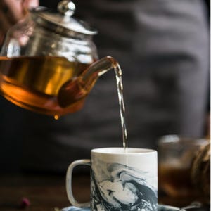 Puede incluir: Una tetera de vidrio vierte té ámbar en una taza blanca con un diseño de mármol gris. La taza está sobre un platillo a juego. La escena evoca un momento de relax con té.