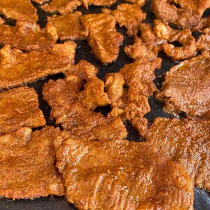 May include: Close-up of a pan filled with thin slices of seasoned and browned meat. The meat is arranged in a random pattern, with some pieces overlapping.