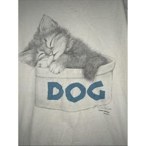 May include: A white t-shirt featuring a pencil-drawn illustration of a sleeping gray kitten nestled in a container labeled "DOG" in blue letters. The artwork includes the artist's signature and a copyright notice.