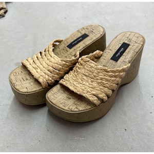 May include: A pair of platform sandals with woven raffia straps and cork-style footbeds. The sandals have a square toe and a wedge heel. The brand name "aldor alian" is printed on the footbed.
