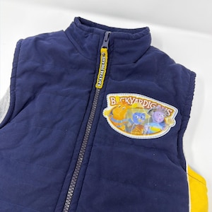 Peut inclure: Gilet matelassé bleu marine avec un écusson "Backyardigans" représentant des personnages de dessins animés. Le gilet a une tirette jaune avec le texte "BACKYARDIGANS" et un panneau latéral gris.