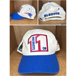Puede incluir: Gorra de béisbol blanca y azul con el número 11 en rojo, la palabra "Patriots" y "QB". La gorra tiene una visera azul y el nombre "BLEDSOE" en la parte posterior. El logotipo de los Patriots está en el lateral.
