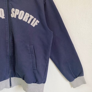 May include: A navy blue zip-up jacket with a gray ribbed cuff and waistband. The jacket has a pocket on the left side and the word "SPORTIF" in gray lettering on the front.