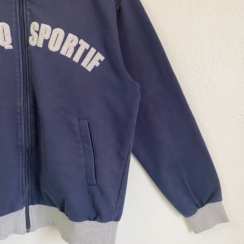 May include: A navy blue zip-up jacket with a gray ribbed cuff and waistband. The jacket has a pocket on the left side and the word "SPORTIF" in gray lettering on the front.