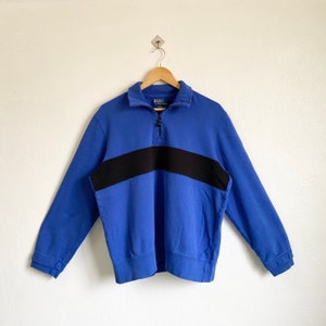 May include: A blue 1/4 zip sweatshirt with a black stripe across the chest. The sweatshirt has a collared neckline and long sleeves.