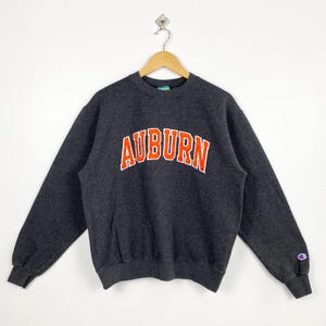 May include: Dark gray Champion sweatshirt with the word "AUBURN" in orange block letters with a white outline. The sweatshirt has a crew neck and long sleeves. A small Champion logo is on the left sleeve.