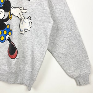 K&ouml;nnte beinhalten: Ein graues Sweatshirt mit einem Cartoon-Bild von Minnie Mouse, die ein rotes Kleid mit wei&szlig;en Punkten tr&auml;gt und eine wei&szlig;e Tasche h&auml;lt. Das Sweatshirt hat einen gerippten Rundhalsausschnitt und B&uuml;ndchen.
