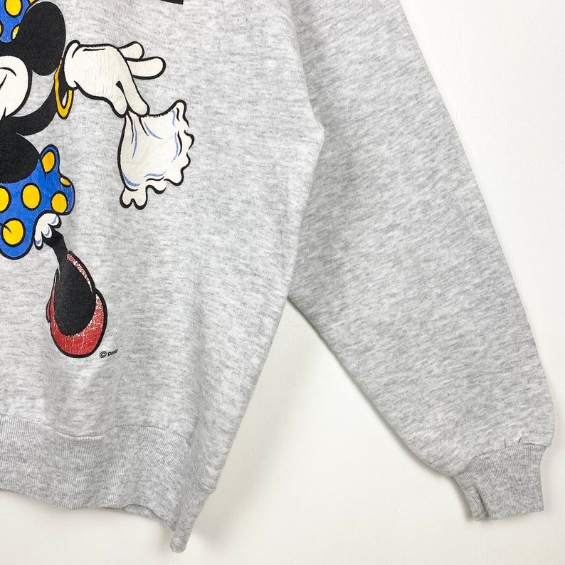 K&ouml;nnte beinhalten: Ein graues Sweatshirt mit einem Cartoon-Bild von Minnie Mouse, die ein rotes Kleid mit wei&szlig;en Punkten tr&auml;gt und eine wei&szlig;e Tasche h&auml;lt. Das Sweatshirt hat einen gerippten Rundhalsausschnitt und B&uuml;ndchen.