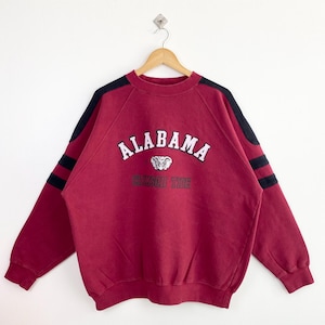 May include: A red sweatshirt with black stripes on the sleeves. The sweatshirt has the words "ALABAMA" and "Crimson Tide" printed on the front in white.