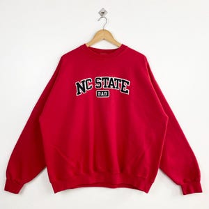 Puede incluir: Sudadera roja con un logotipo bordado en blanco y negro que dice "NC STATE DAD".