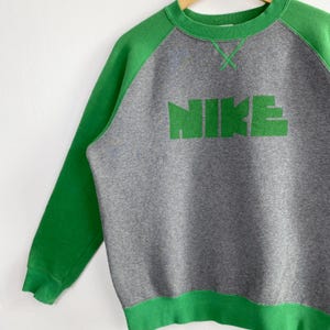 Pu&ograve; includere: Felpa grigia con maniche raglan e finiture verdi. La parola "NIKE" &egrave; stampata a grandi lettere verdi sul petto. La felpa &egrave; realizzata in un materiale morbido e ha uno scollo tondo.