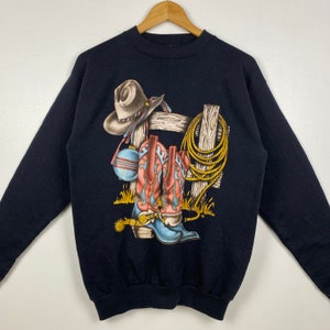 May include: Black sweatshirt with a graphic print of cowboy boots, a cowboy hat, and a rope hanging on a wooden fence.