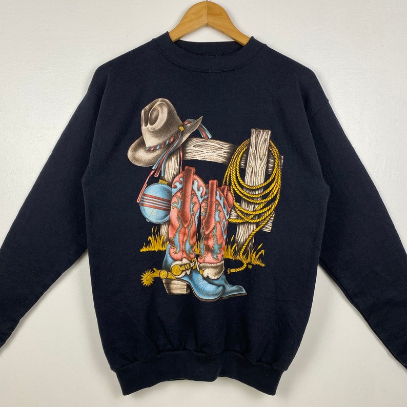 May include: Black sweatshirt with a graphic print of cowboy boots, a cowboy hat, and a rope hanging on a wooden fence.