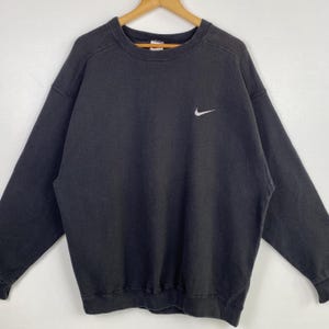 Puede incluir: Una sudadera Nike negra con un logotipo Swoosh blanco en el pecho izquierdo. La sudadera tiene cuello redondo y mangas largas.