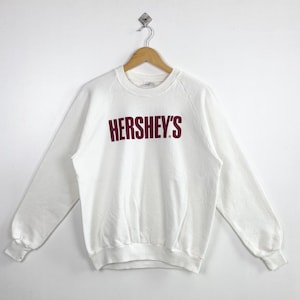 May include: A white sweatshirt with the word "HERSHEY'S" printed in maroon across the chest. The long-sleeved top is hanging on a wooden hanger against a white background. The sweatshirt has a crew neck and ribbed cuffs.