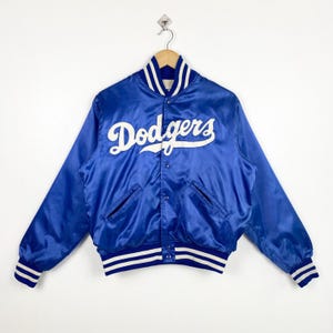 Può includere: Una giacca da baseball in satin blu con bordature bianche a righe sui polsini e in vita. La giacca presenta un logo "Dodgers" ricamato in bianco sul davanti.