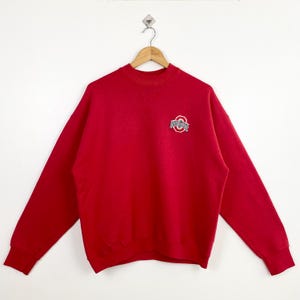 Può includere: Felpa rossa con un logo Ohio State bianco e grigio sul davanti.
