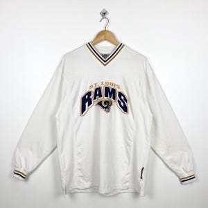 Puede incluir: Sudadera blanca de manga larga con un logotipo bordado de los St. Louis Rams en azul marino y dorado. La sudadera tiene cuello en V y puños de canalé.
