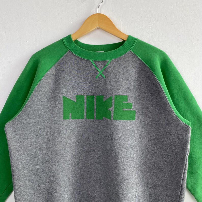 Pu&ograve; includere: Una felpa grigia con maniche e colletto verdi. La parola "NIKE" &egrave; stampata a grandi lettere verdi sul petto. La felpa &egrave; appesa a una gruccia di legno.