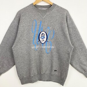 Puede incluir: Sudadera gris con un logotipo azul y blanco de la Universidad de Carolina del Norte con una mascota de dibujos animados. El logotipo dice "University of North Carolina" y tiene la palabra "Rames" en la parte inferior.