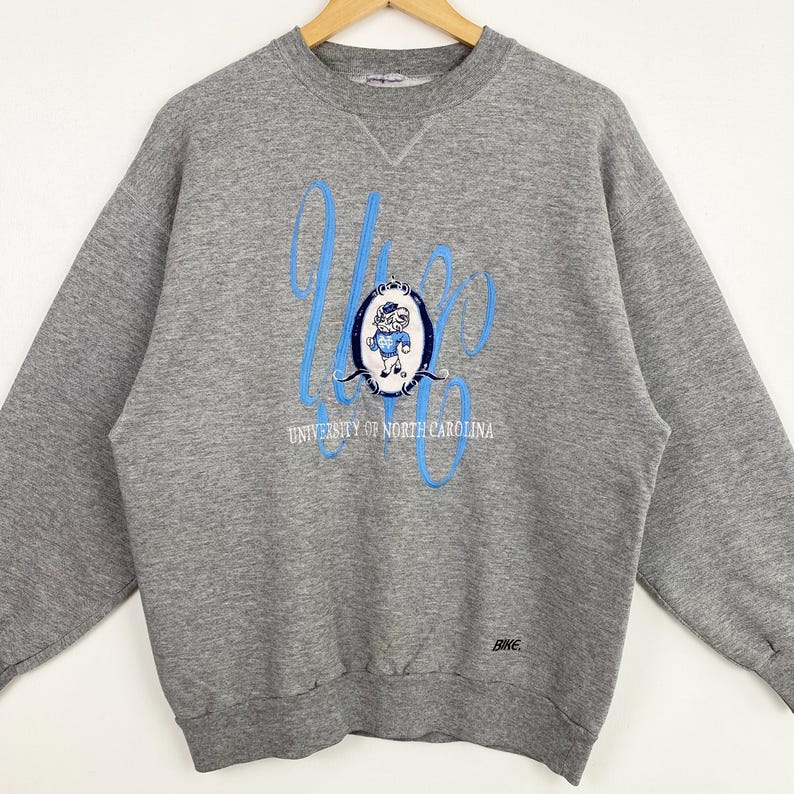Puede incluir: Sudadera gris con un logotipo azul y blanco de la Universidad de Carolina del Norte con una mascota de dibujos animados. El logotipo dice "University of North Carolina" y tiene la palabra "Rames" en la parte inferior.