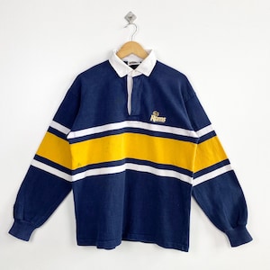 Peut inclure: Un maillot de rugby bleu marine à manches longues avec des rayures blanches et jaunes. Le maillot a un col blanc et un petit logo brodé qui dit "Rams". Le maillot est suspendu à un cintre en bois.