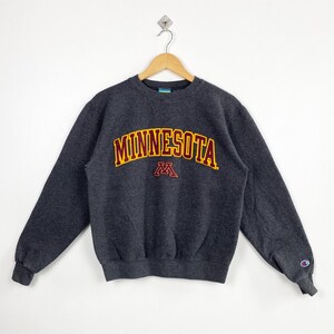 May include: A dark gray sweatshirt with the word "MINNESOTA" in gold and maroon lettering across the chest. Below the word is a maroon "M" logo. The sweatshirt has long sleeves and a crew neck.