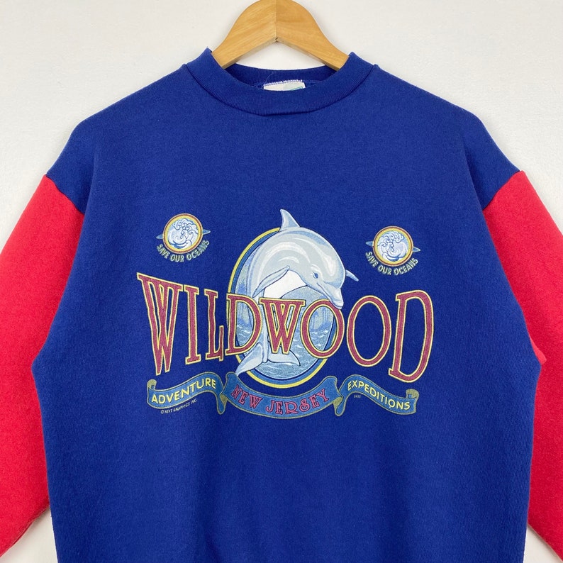 May include: Blue and red sweatshirt with a dolphin jumping out of a circle with the text "Wildwood, New Jersey, Adventure Expeditions" and "Save Our Oceans" on either side.