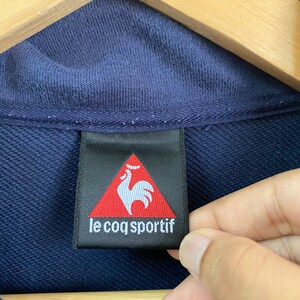 May include: A navy blue clothing label with a red triangle containing a white rooster and the text "le coq sportif".