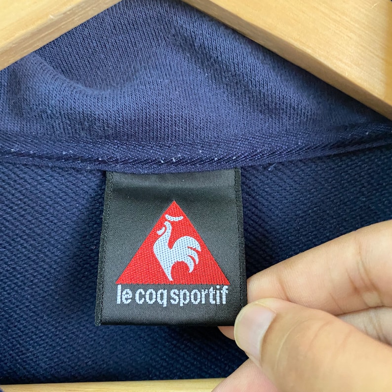 May include: A navy blue clothing label with a red triangle containing a white rooster and the text "le coq sportif".