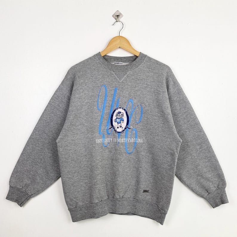 Puede incluir: Sudadera gris con un logotipo azul y blanco de la Universidad de Carolina del Norte y el texto "UNC" en una fuente estilizada.