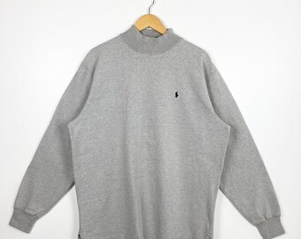 May include: A heather gray long-sleeve sweatshirt with a mock neck collar. The sweatshirt features a small black logo on the chest. The garment is hanging on a wooden hanger against a white background.