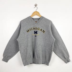 Könnte beinhalten: Grauer Sweatshirt mit V-Ausschnitt und einem gestickten Logo in Marineblau und Gold, auf dem "Michigan" mit einem "M" in einem Kreis steht.