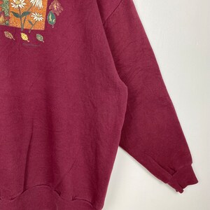 May include: A maroon sweatshirt with a floral print on the front. The print features a square frame with flowers and leaves.