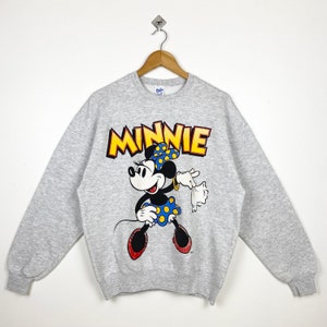 K&ouml;nnte beinhalten: Grauer Pullover mit einem Cartoon-Bild von Minnie Mouse in einem blau-gelb gepunkteten Kleid. Der Text "MINNIE" ist in Gelb &uuml;ber ihr gedruckt.